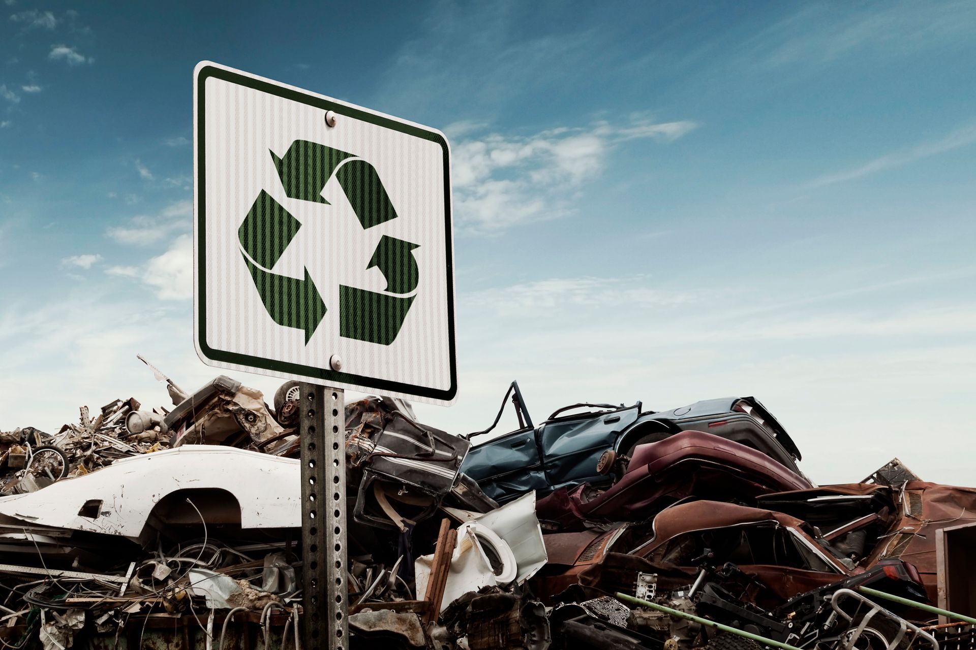 A green recycling sign stands before a pile of crushed cars and scrap metal at a junkyard.