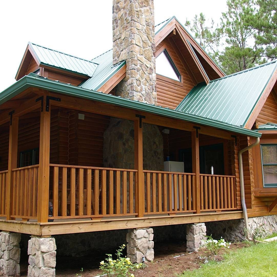 Wooden cabin with green metal roof, stone chimney, and covered porch in a wooded setting