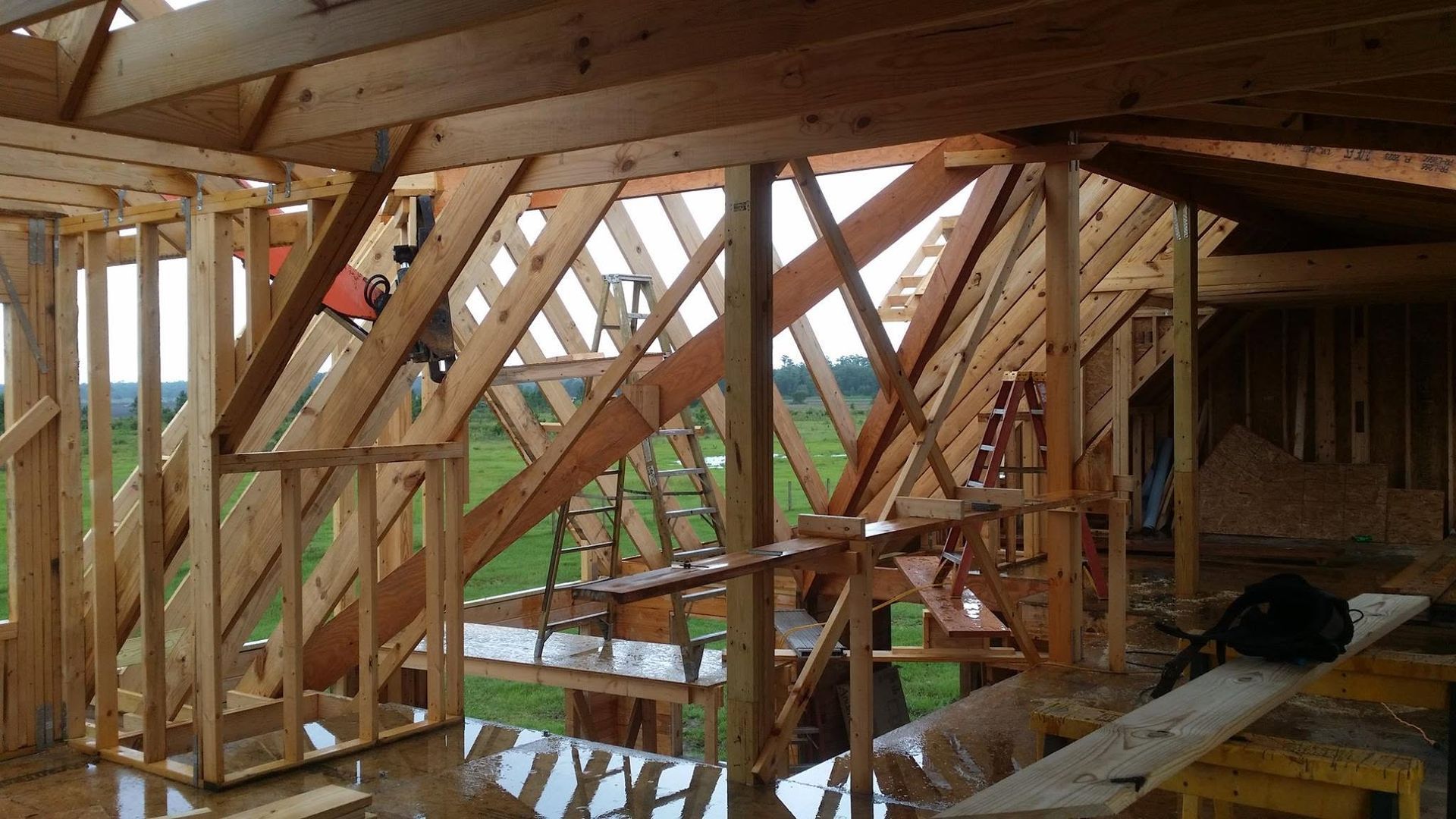 Wood-framed house under construction with exposed beams and a wet floor, seen through open wall framing
