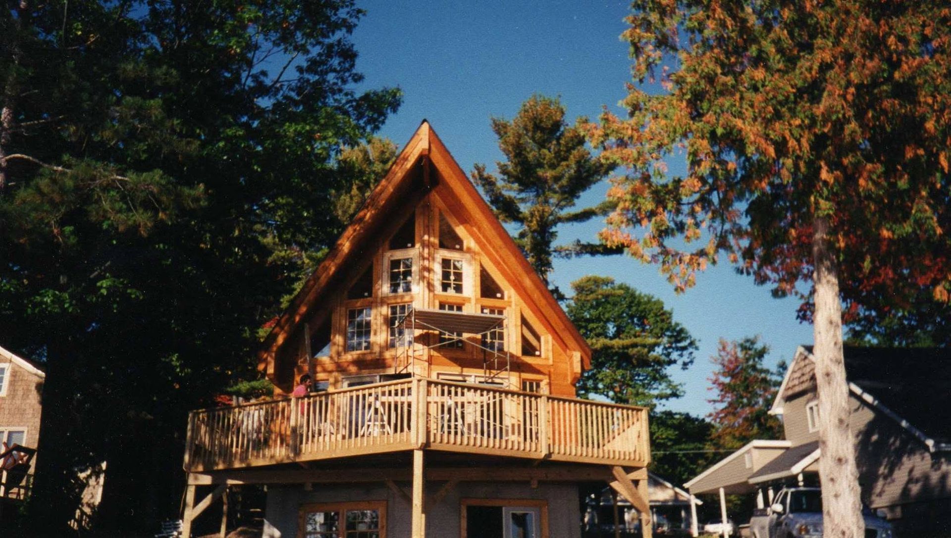 A wooden chalet-style house with a steep roof and wraparound porch among tall trees on a sunny day