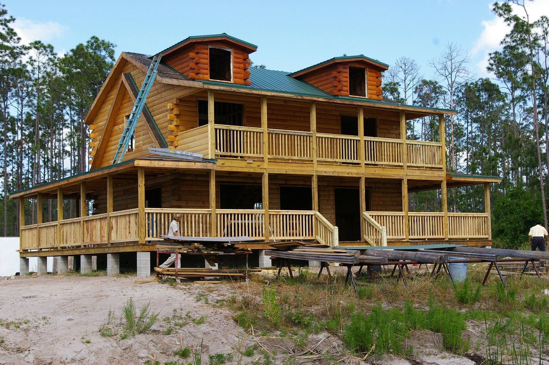 Large two-story wooden lodge under construction, with a wraparound porch and trees in the background