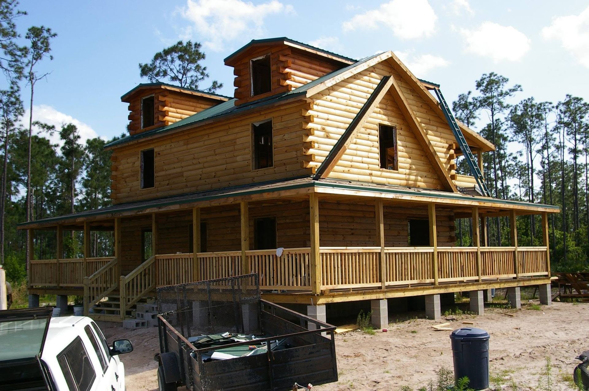 Wooden cabin under construction with a wraparound porch, surrounded by trees and building materials.