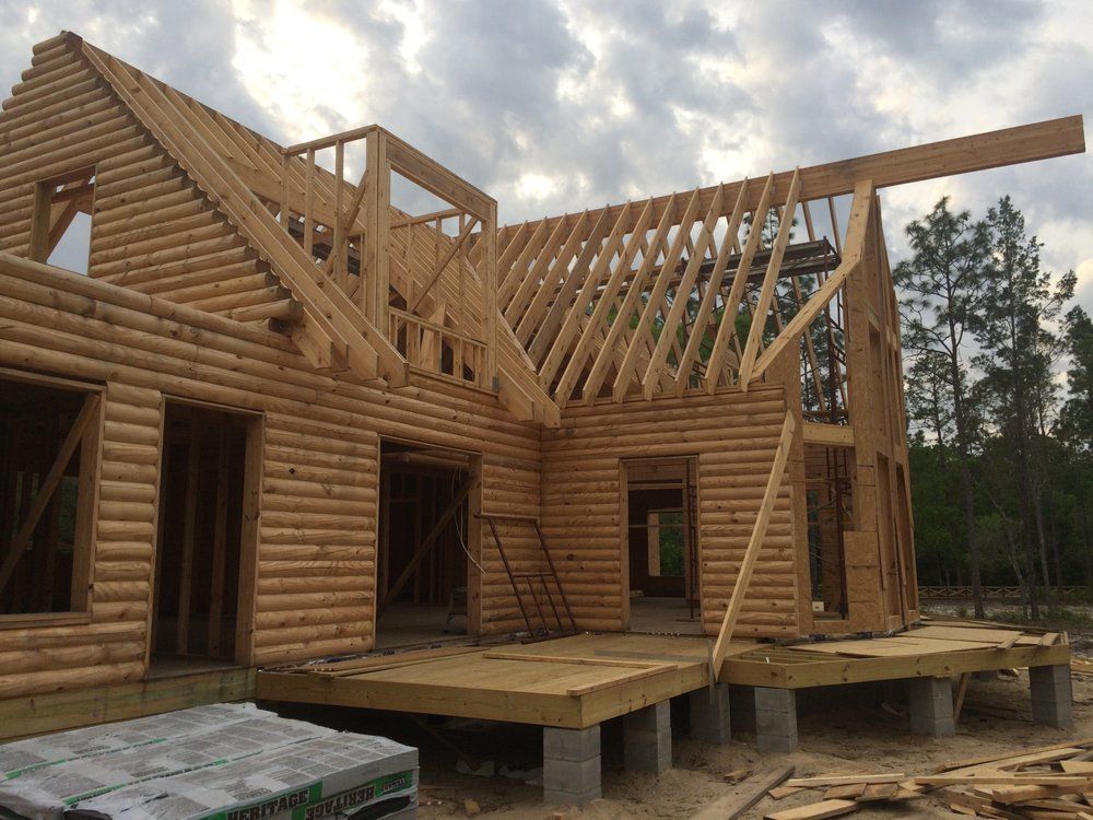 Partially built log house with exposed roof framing on a raised foundation in a wooded area