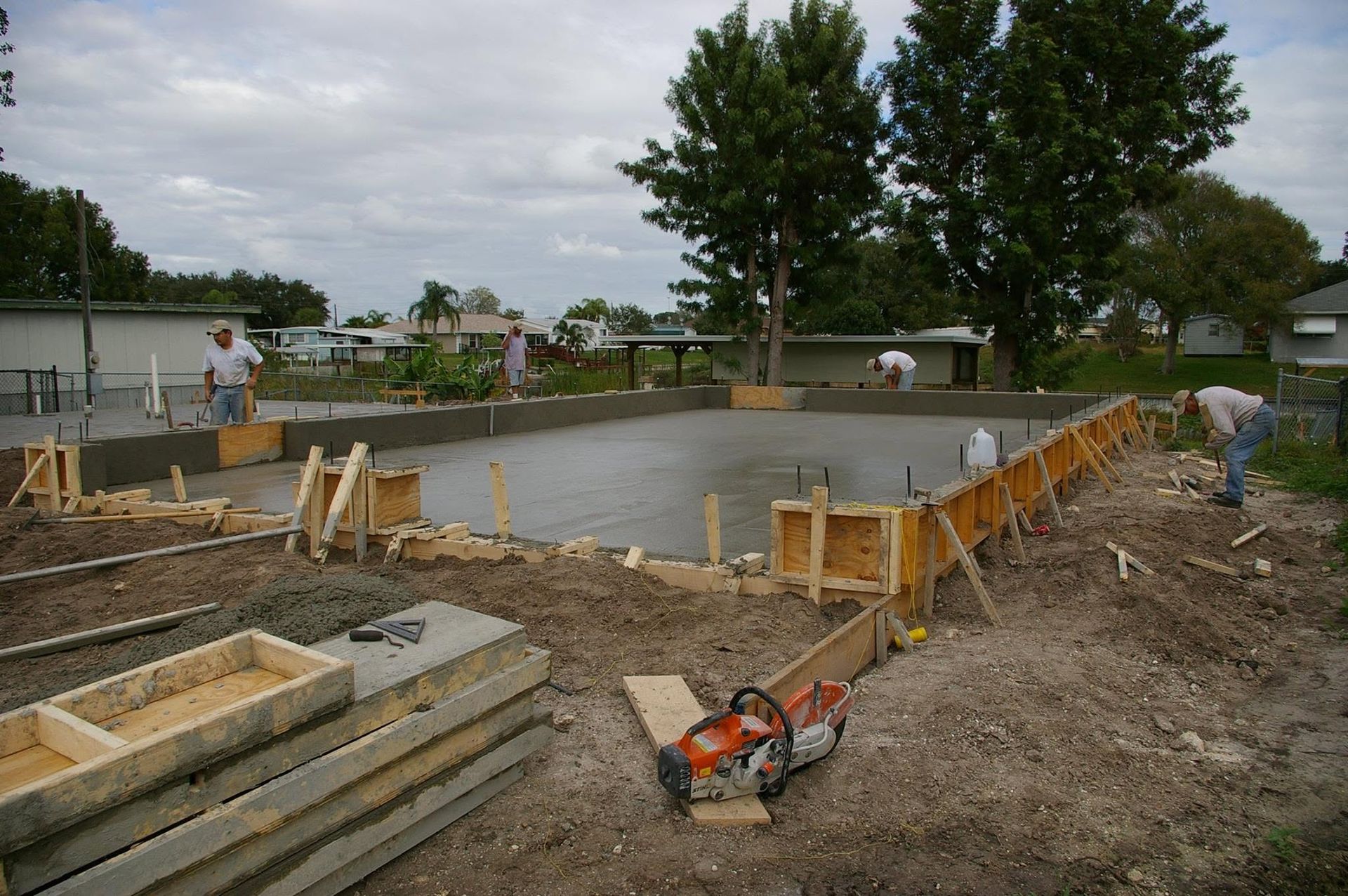 Concrete foundation under construction with wooden forms, dirt, tools, and trees in the background