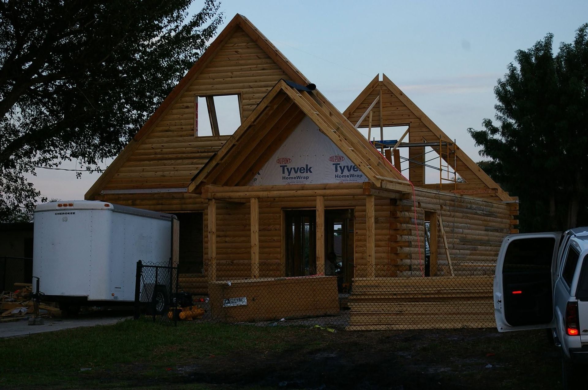 Partially built wooden house under construction at dusk, with trailer and van beside it