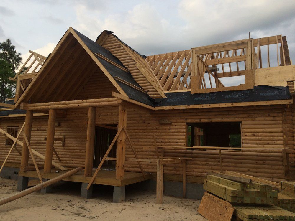 Wooden cabin under construction with exposed roof framing and stacked lumber nearby