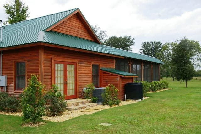 Wooden cabin with green metal roof on a grassy lawn, surrounded by trees.