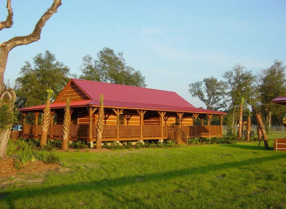 Wooden lodge with a red roof in a grassy rural setting, surrounded by trees under a blue sky