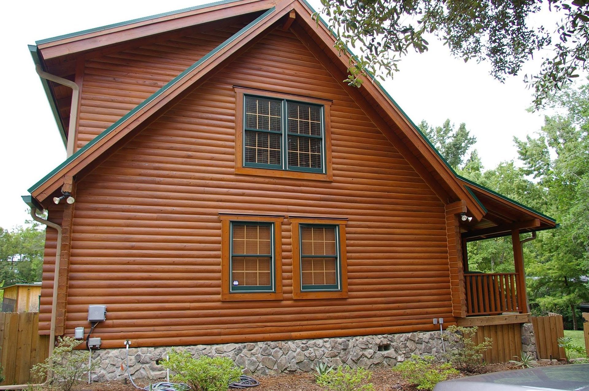 Side view of a rustic wooden house with a gabled roof and windows, surrounded by trees and landscaping