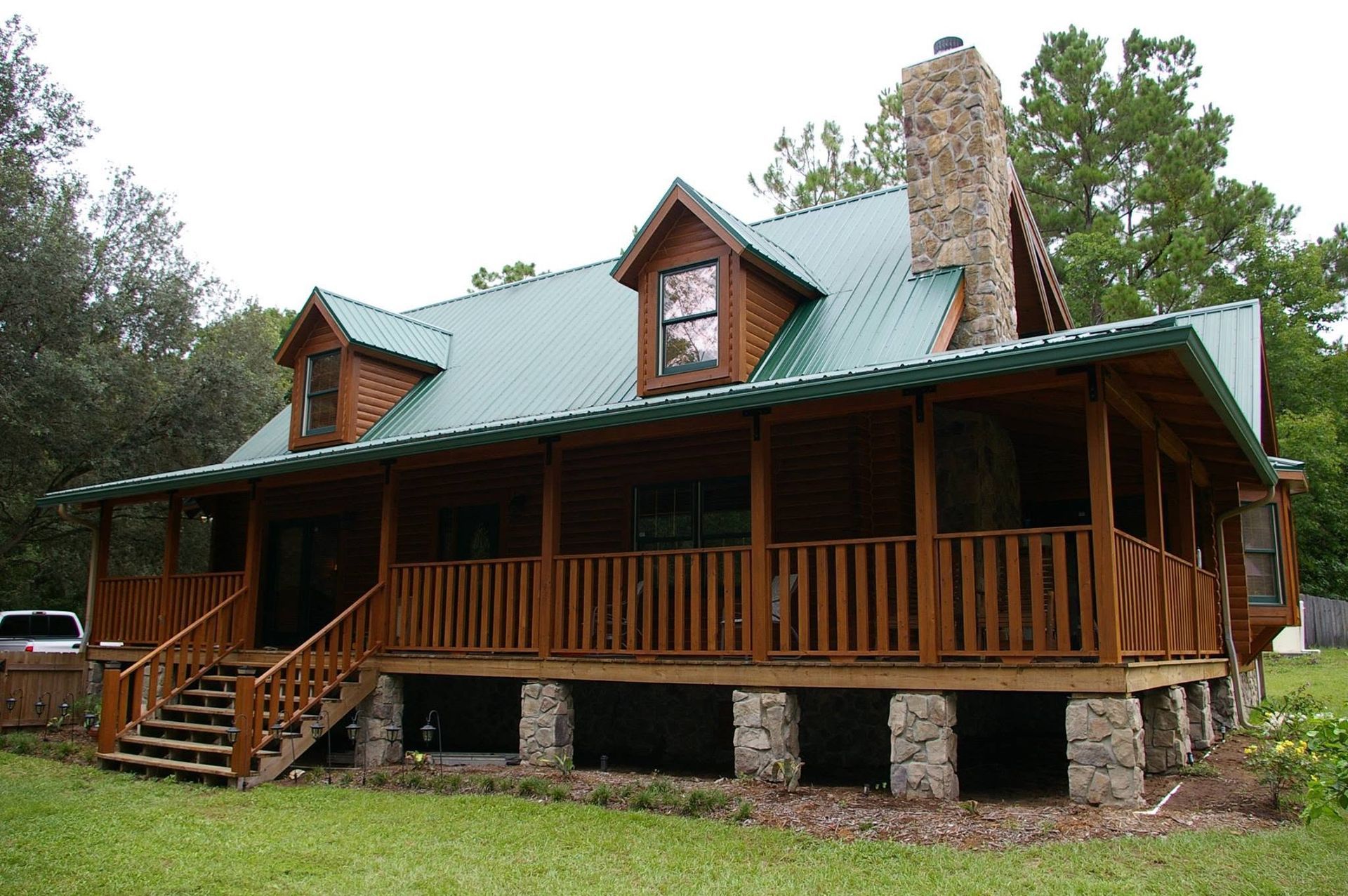 Log cabin with green metal roof, covered porch, stone chimney, and front steps in a wooded setting