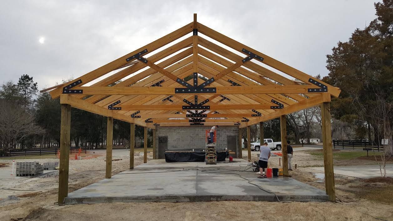 Unfinished open-sided pavilion with exposed wooden roof trusses on a concrete slab at a construction site