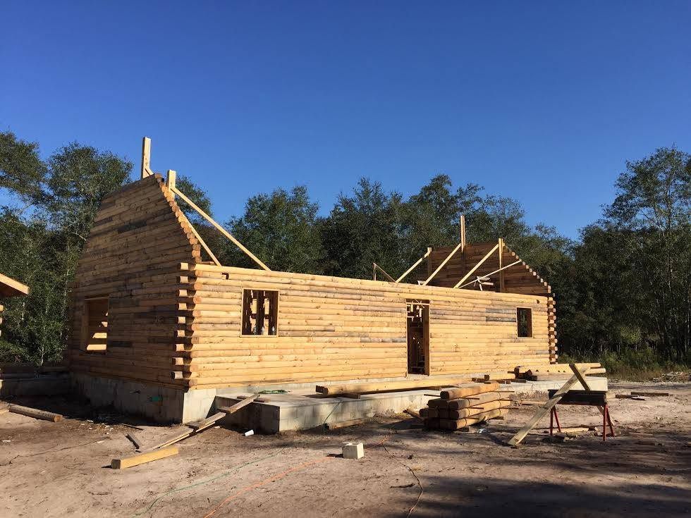 Partially built wooden cabin frame on a dirt lot under a clear blue sky