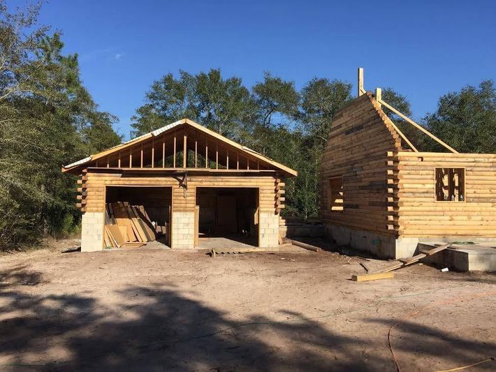 Two unfinished wooden buildings under construction on a dirt lot, with trees and blue sky in the background