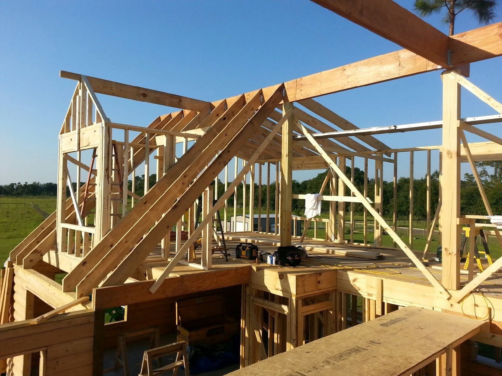 Wood-framed house under construction with roof trusses and open walls in a grassy field