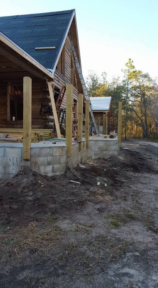 Wooden house under construction with stone foundation, posts, and a dirt yard at sunset.