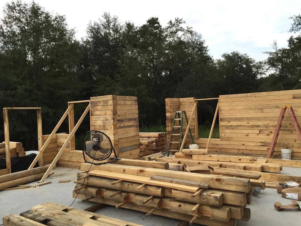 Outdoor construction site with stacked lumber, wooden walls, ladders, and tools on a concrete slab