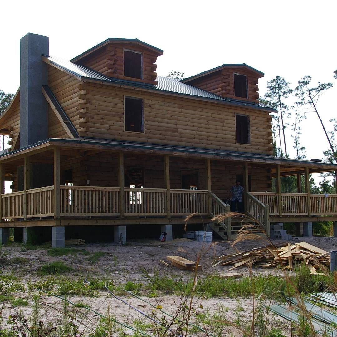 Large rustic wooden cabin with a covered porch, set in a clearing with scattered lumber and workers nearby