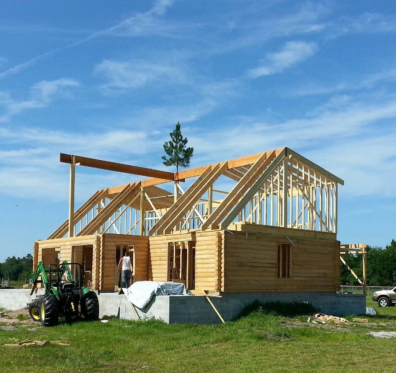 Partially built wooden house with roof framing on a grassy site under a blue sky