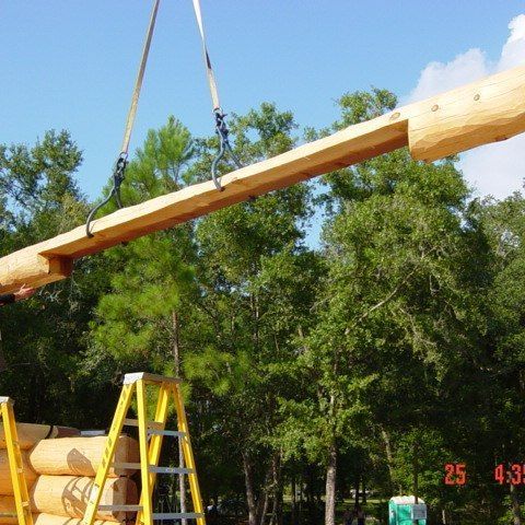 Large wooden beam being lifted by a crane above a ladder in a wooded outdoor area