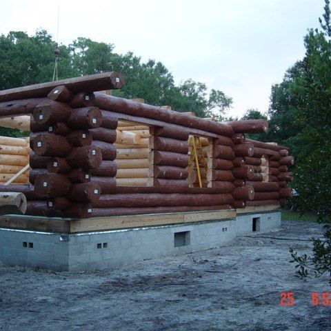 Log cabin under construction on a concrete foundation, with stacked logs and green trees around it.