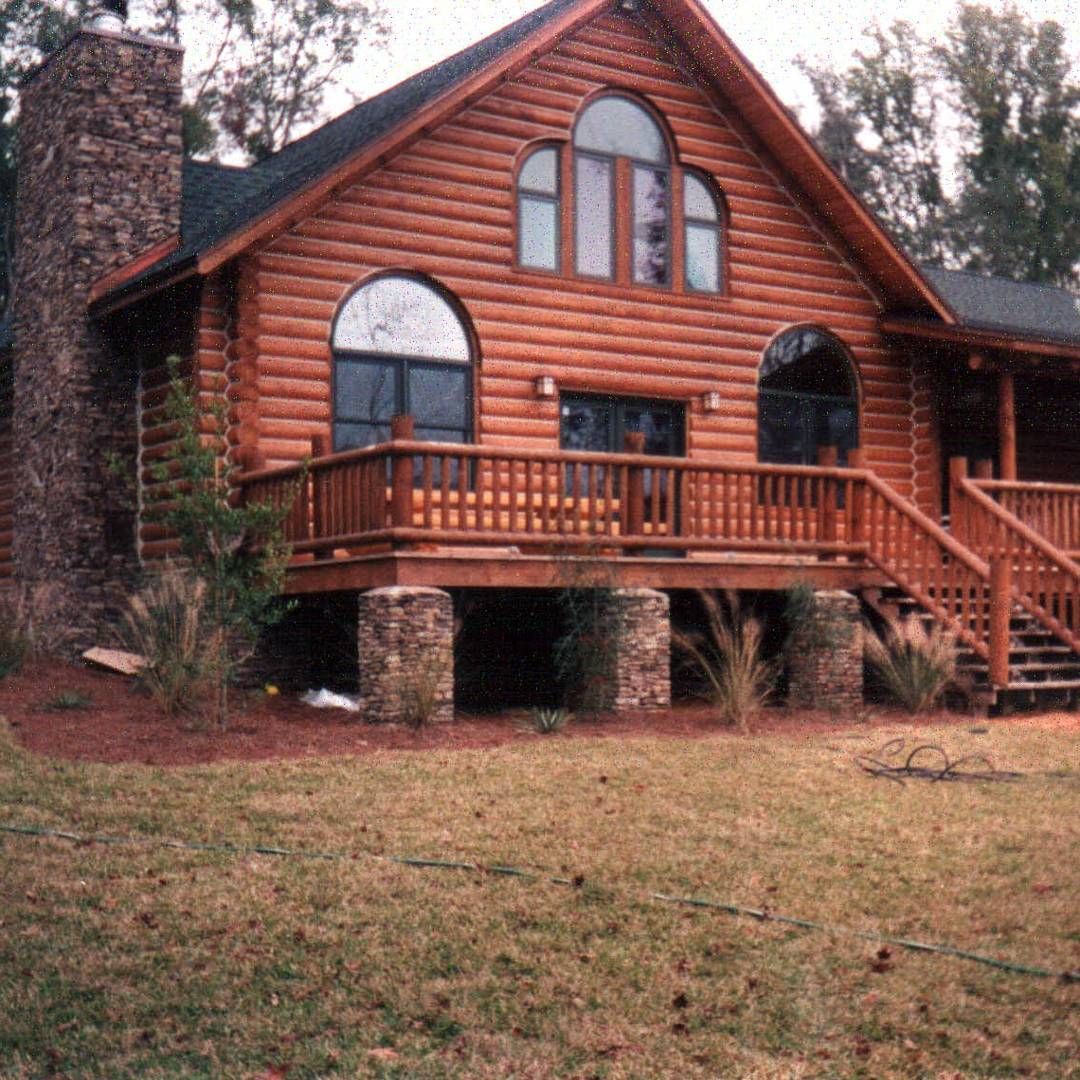 Log cabin with red-brown wood siding, stone chimney, and front porch in a wooded yard