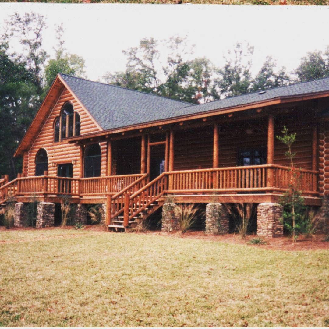 Rustic log cabin with a wooden porch and stone foundation, set in a grassy clearing with trees behind it