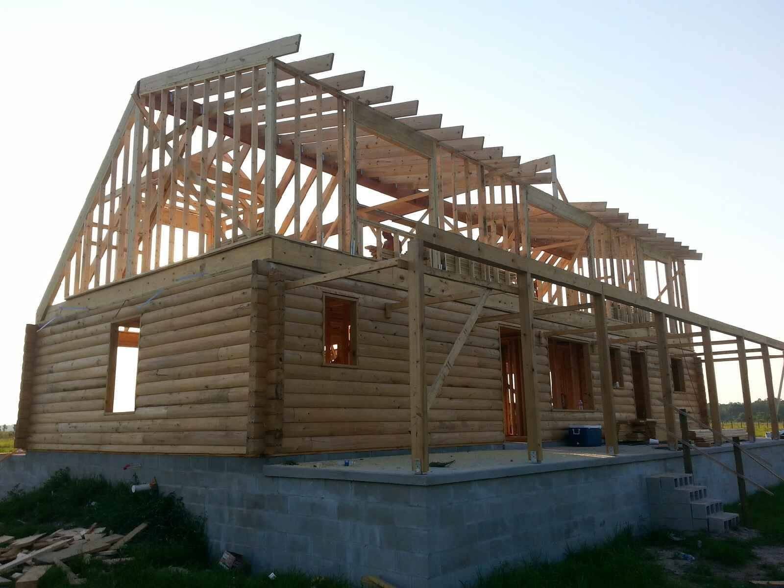 Partially framed wooden house under construction with log walls and roof trusses on a concrete foundation