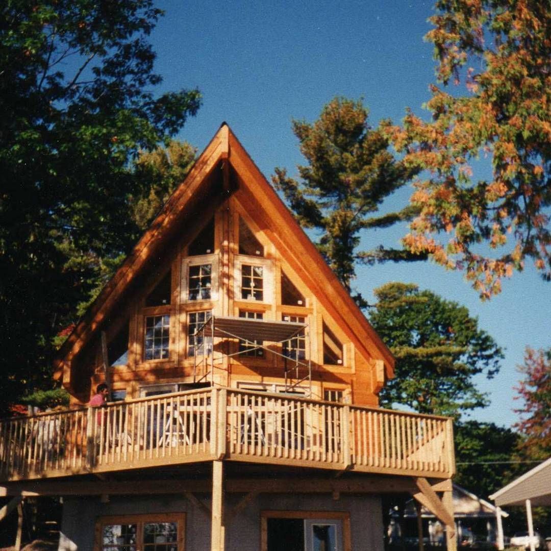 Wooden A-frame cabin with a wraparound deck among tall trees on a sunny day