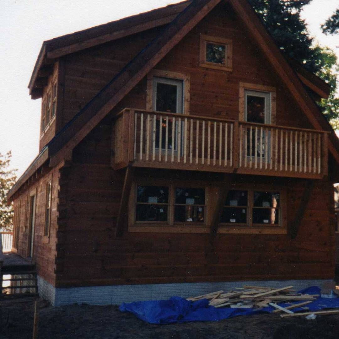 Wooden cabin under construction with a balcony, brown siding, and blue tarp in front.