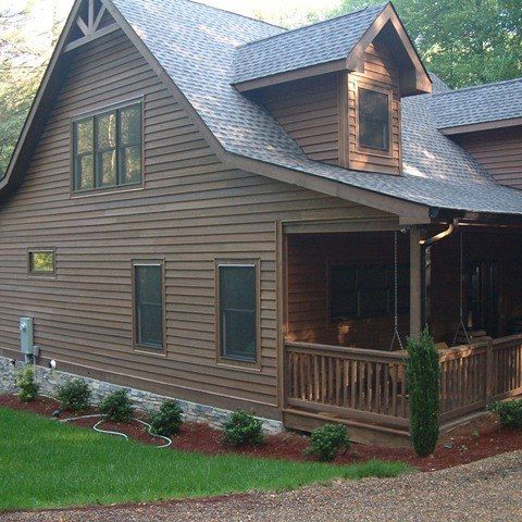 Brown two-story house with a porch, green lawn, and landscaped garden beds