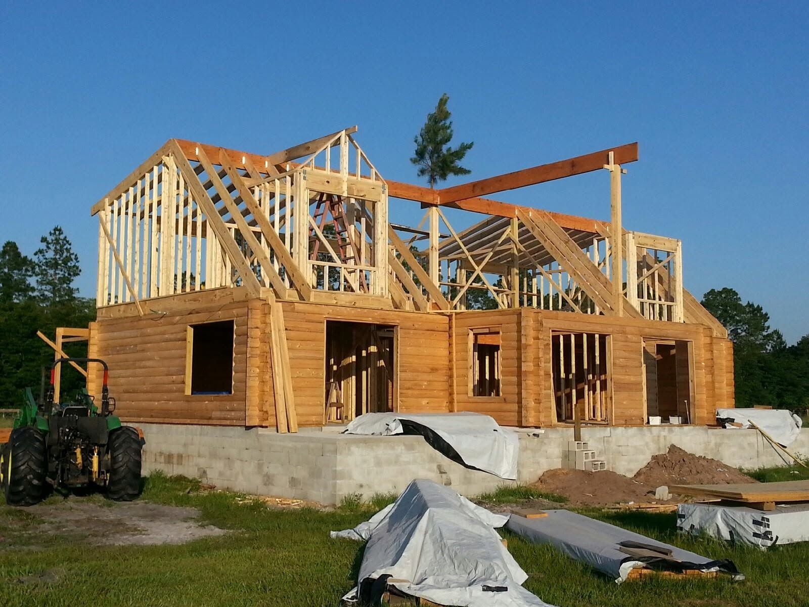 Wood-frame house under construction with roof trusses, concrete foundation, and lumber piles on site