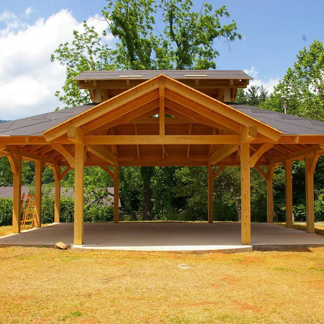 Wooden open-air pavilion with a peaked roof, set in a grassy park under a blue sky.