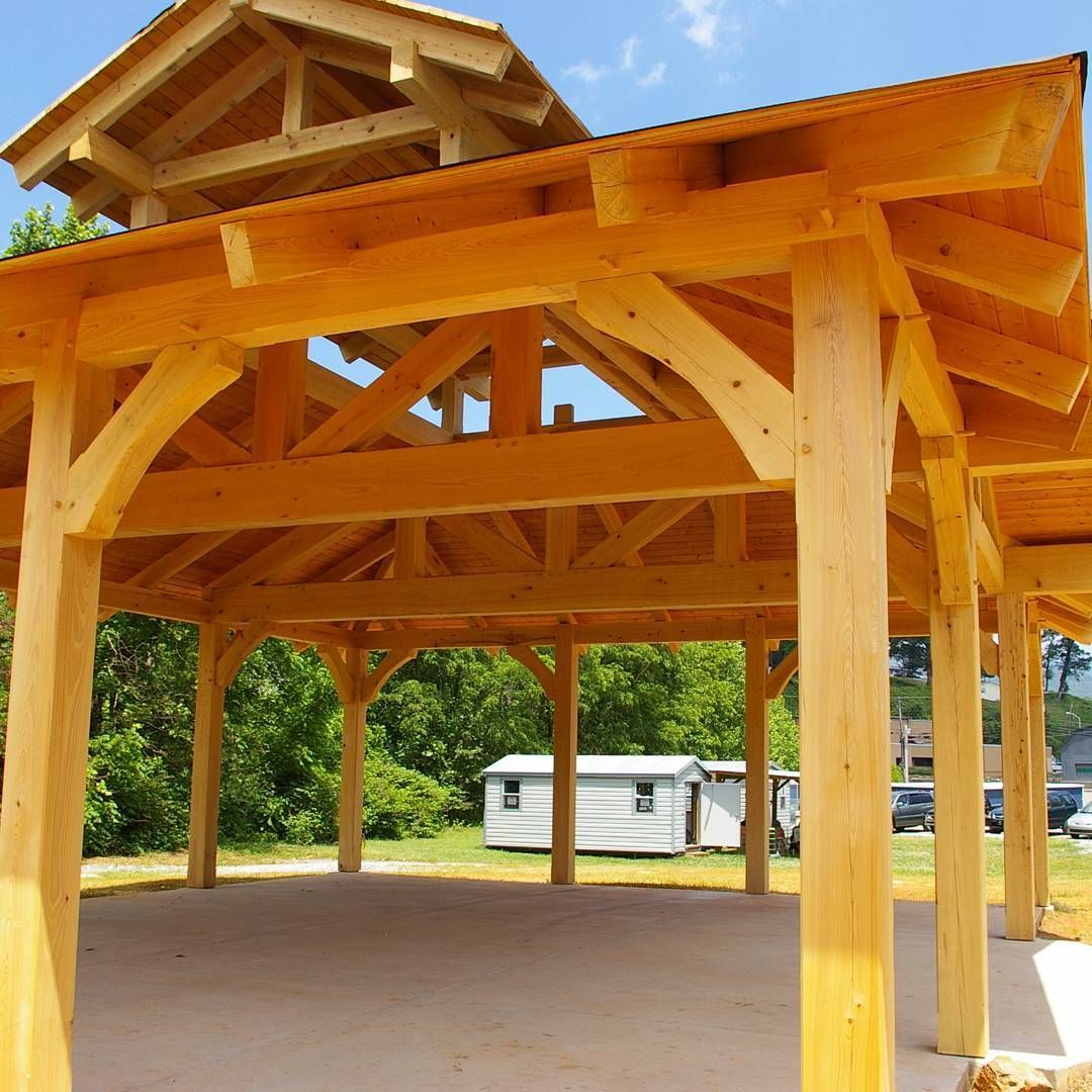 Wooden open-air pavilion with a peaked roof and exposed beams on a concrete pad, surrounded by trees.