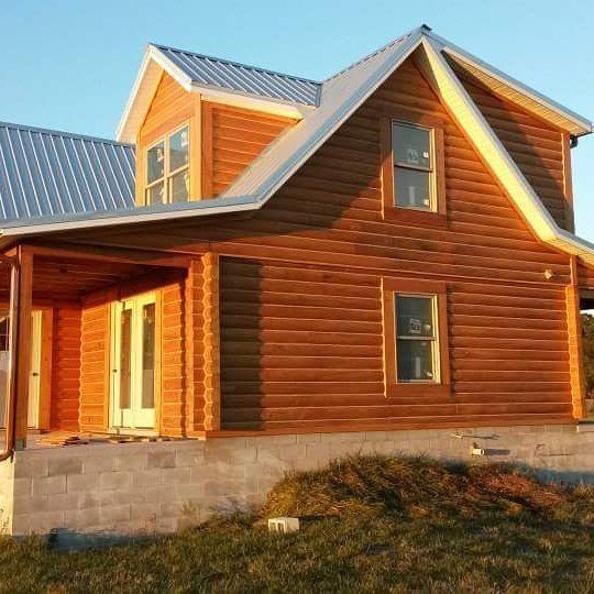Sunlit wooden house with a gray metal roof and stone foundation on a grassy lot