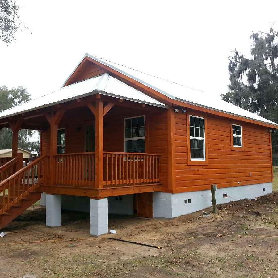Small orange wooden cabin with a front porch and white foundation, set on a dirt lot among trees.