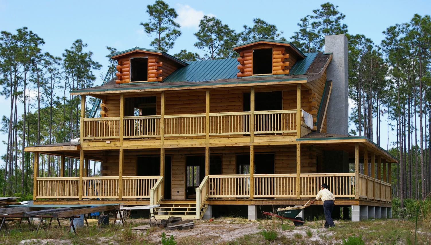 Large unfinished wooden house with wraparound porches and twin dormers in a wooded area