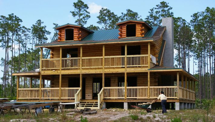 Large unfinished wooden house with wraparound porches and twin dormers in a wooded area