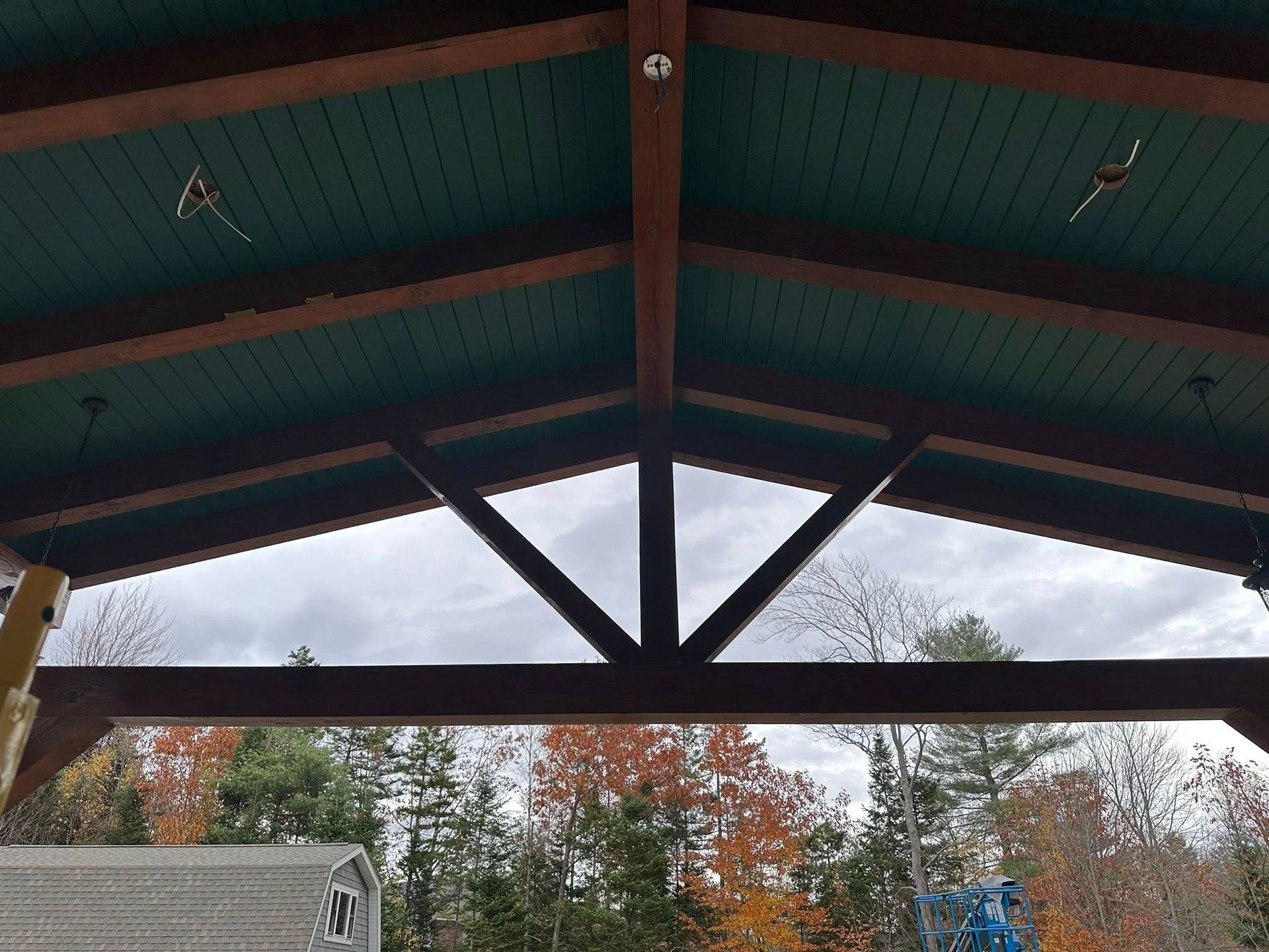 Wooden pavilion roof with teal ceiling, framed by autumn trees and cloudy sky.