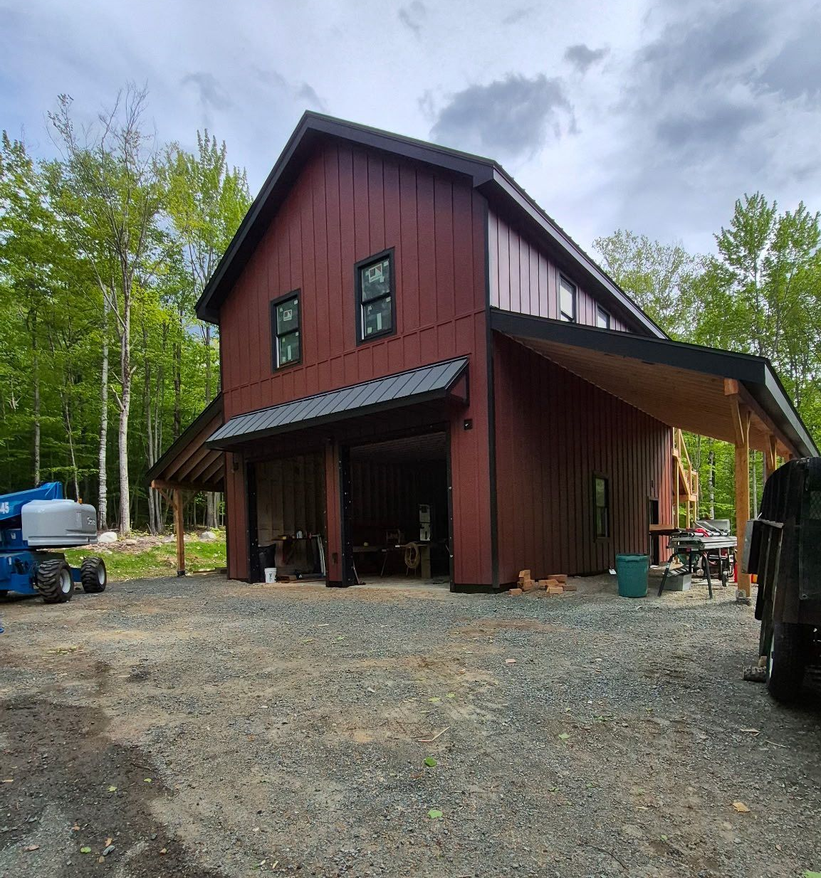 Rustic red barn-style building with black trim and gravel yard, surrounded by trees under a cloudy sky