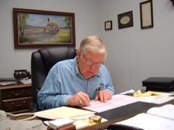 Man reading documents at a desk in an office with papers, framed artwork, and filing cabinets.