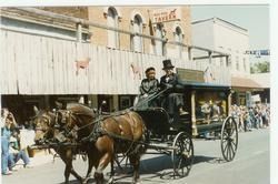 Horse-drawn carriage with two drivers on a city street outside storefronts and spectators