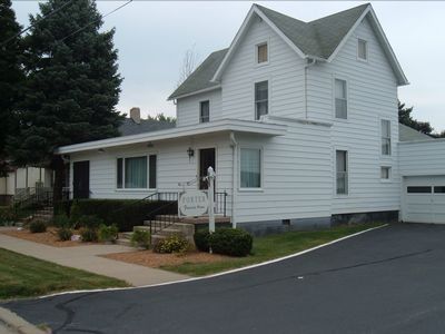 White two-story house with porch, gray roof, and driveway beside a sidewalk