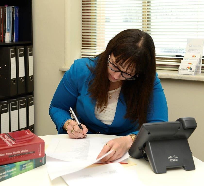 Woman in Blue Jacket Writing at a Desk With Phone and Books — Umbrella Legal in Cardiff, NSW
