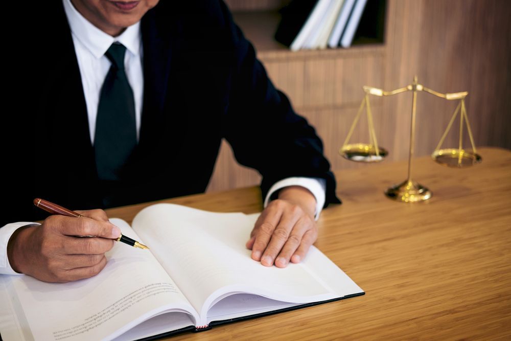 Lawyer in Suit Writing in Large Book; Scales of Justice on Desk — Umbrella Legal in Wyong, NSW