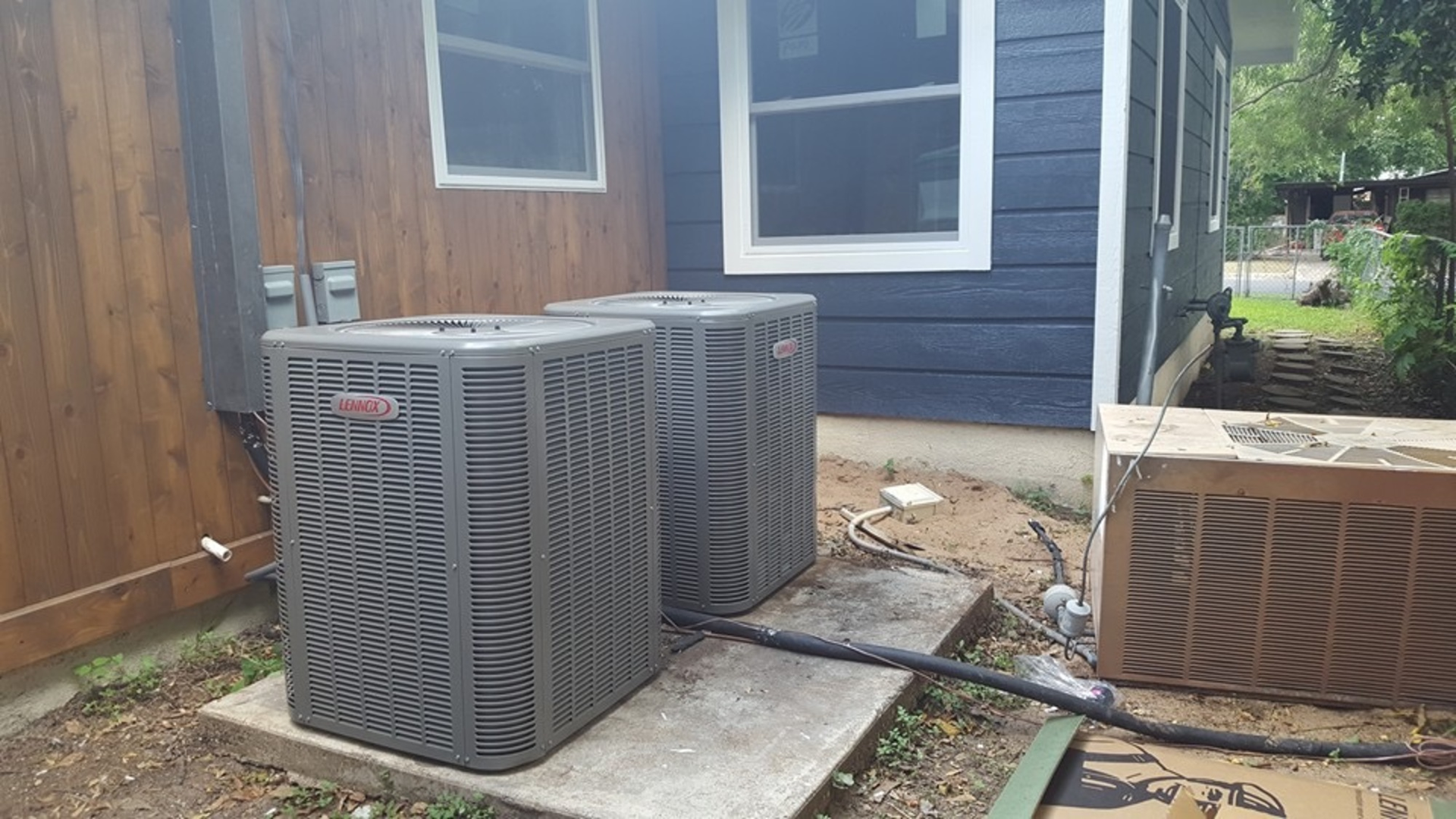 Two air conditioners are sitting in front of a blue house.