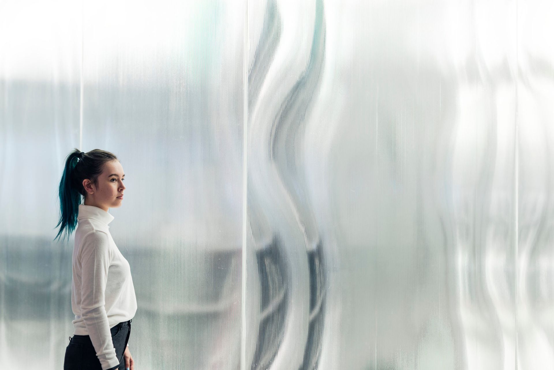 A person with a dark ponytail wearing a white turtleneck stands in profile, gazing at a tall, wavy, translucent wall.