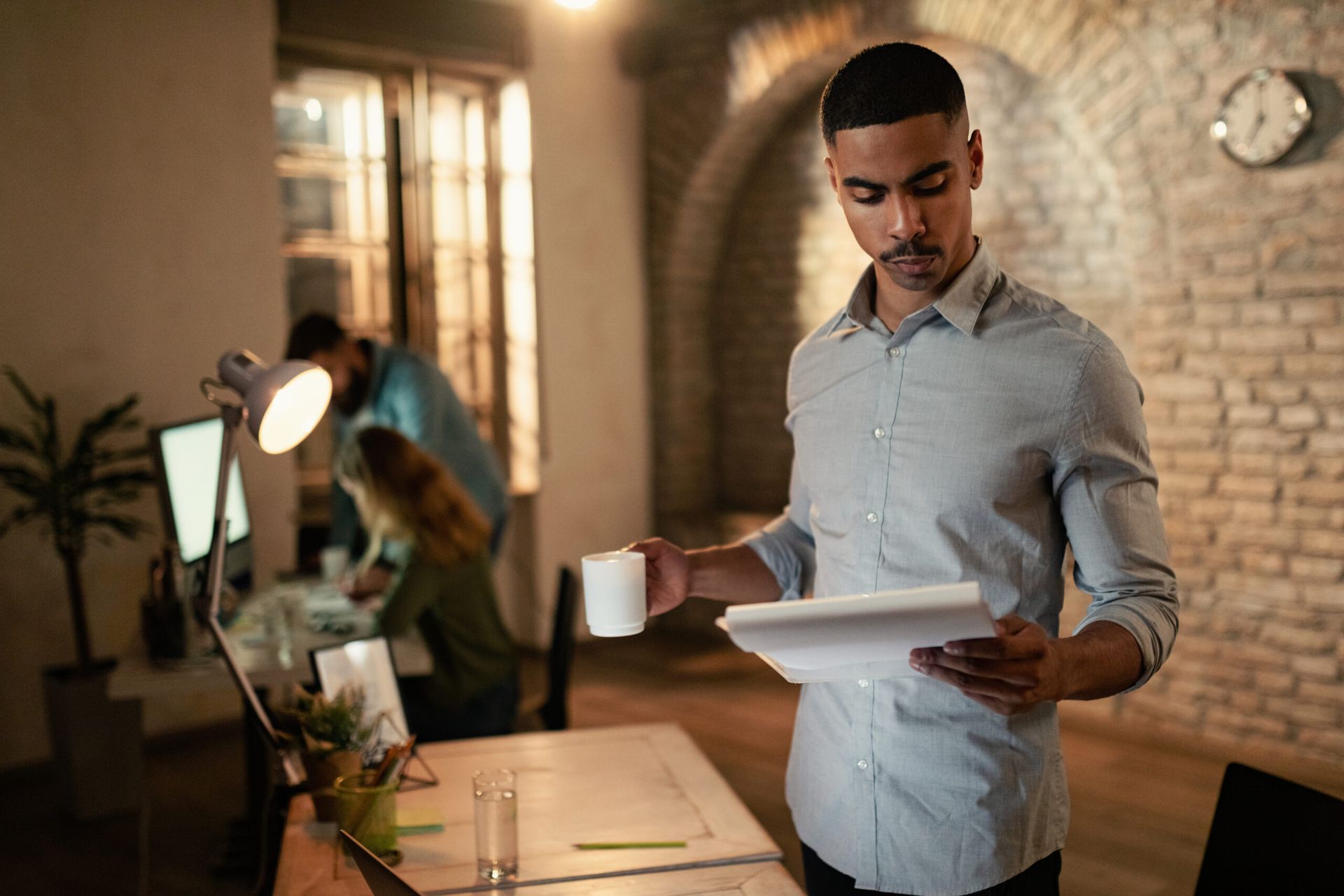 Man holding a coffee mug and plate in a warmly lit office with a desk, lamp, and laptop in the background