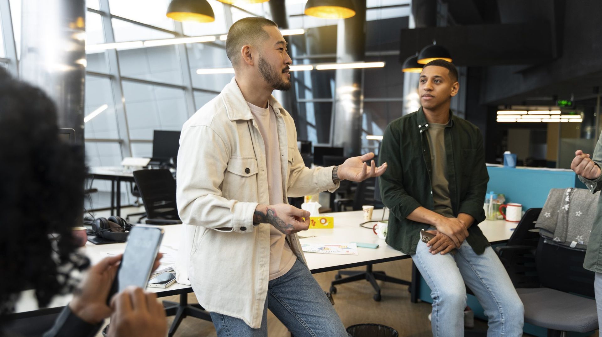 Two people having a discussion in a modern office, one standing and gesturing while the other sits on a desk.