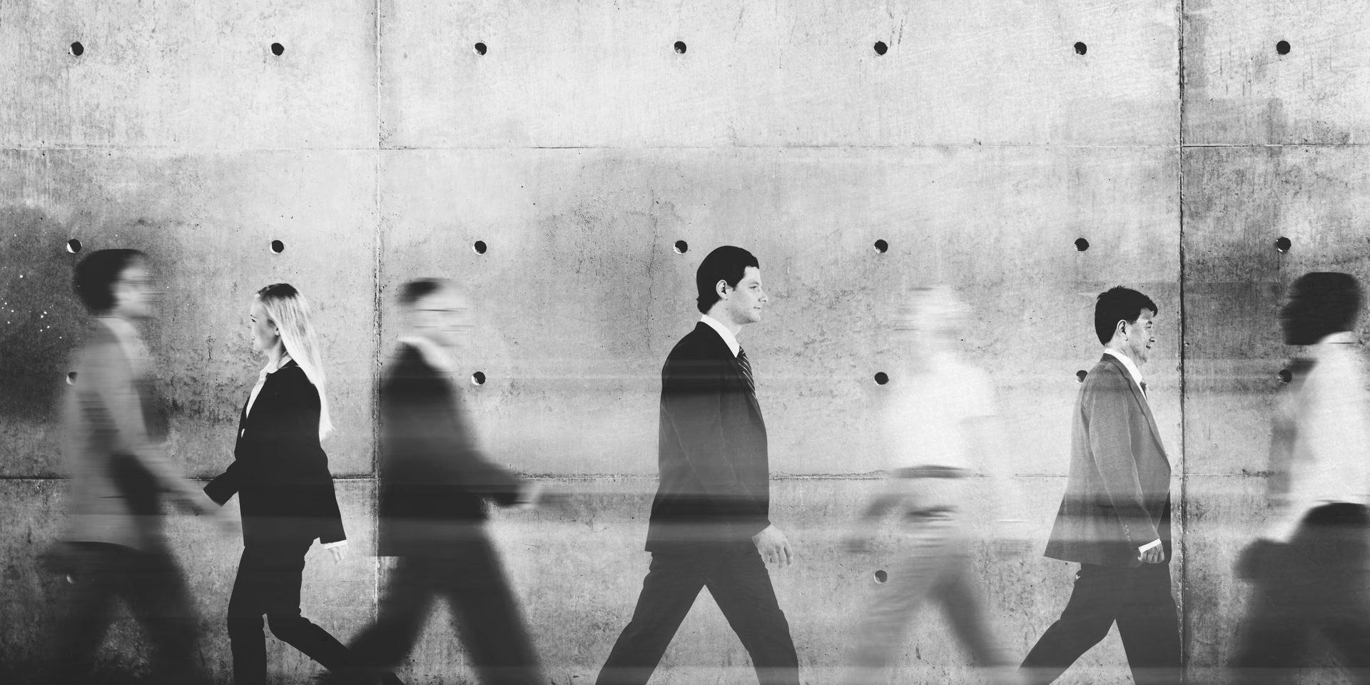 Blurry black-and-white silhouettes of people walking left to right along a concrete wall.