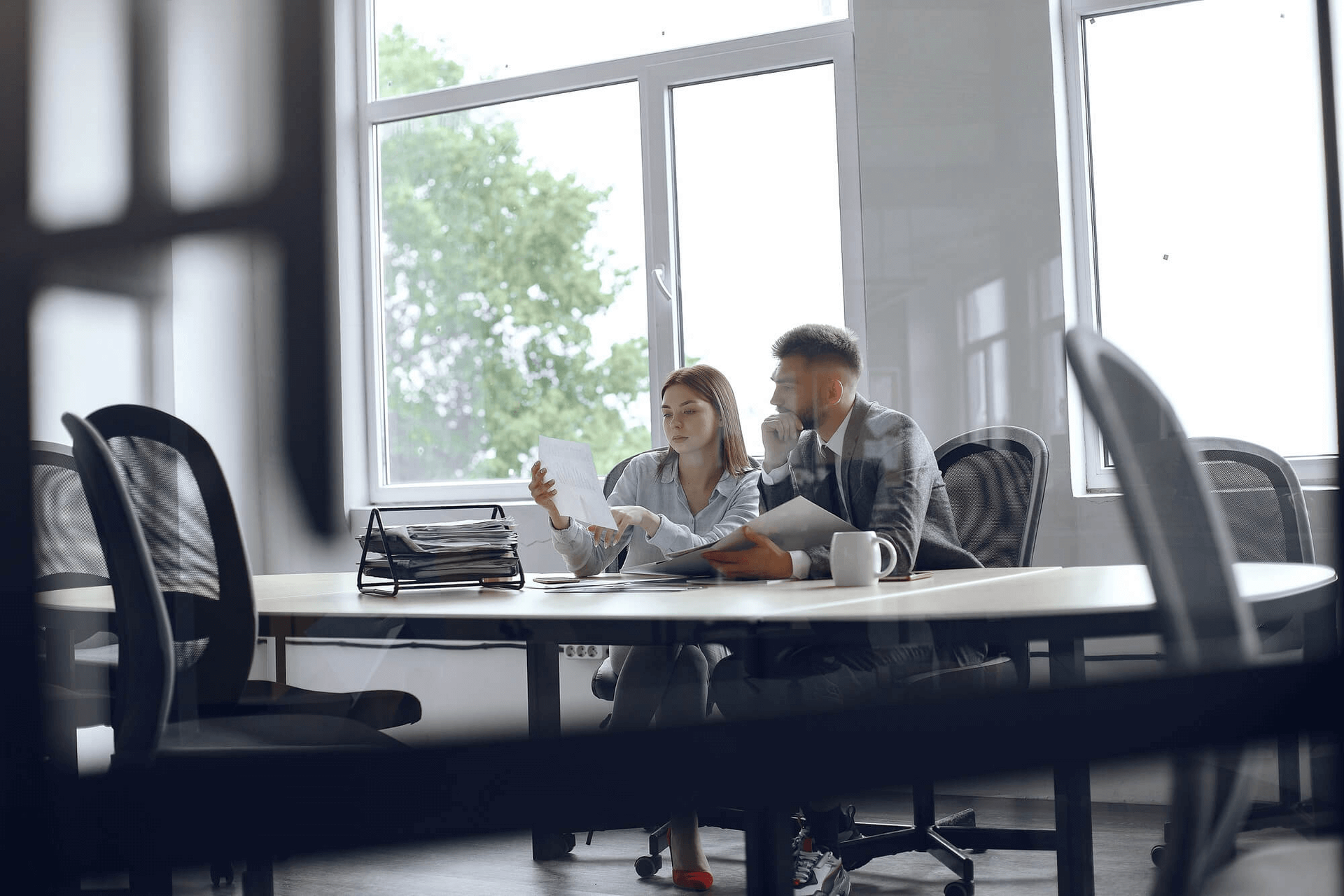 Two coworkers talking at a conference table in a bright office with large windows